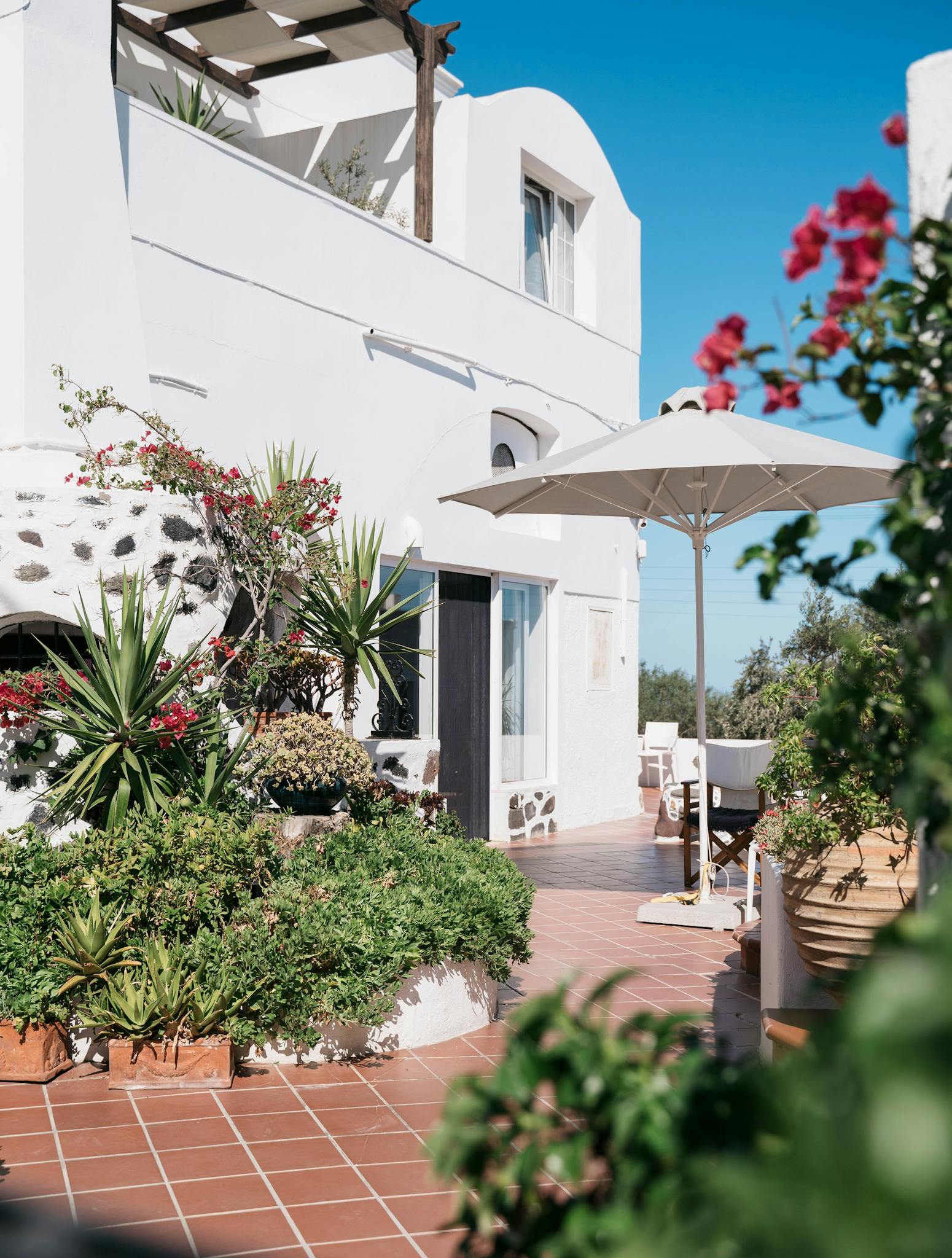Sunlit Santorini villa with white architecture and vibrant garden, perfect for relaxing outdoors.